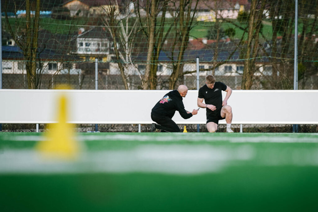 Im Hintergrund einer Outdoorfläche sind Personal Trainer und Kunde im Gespräch zu sehen