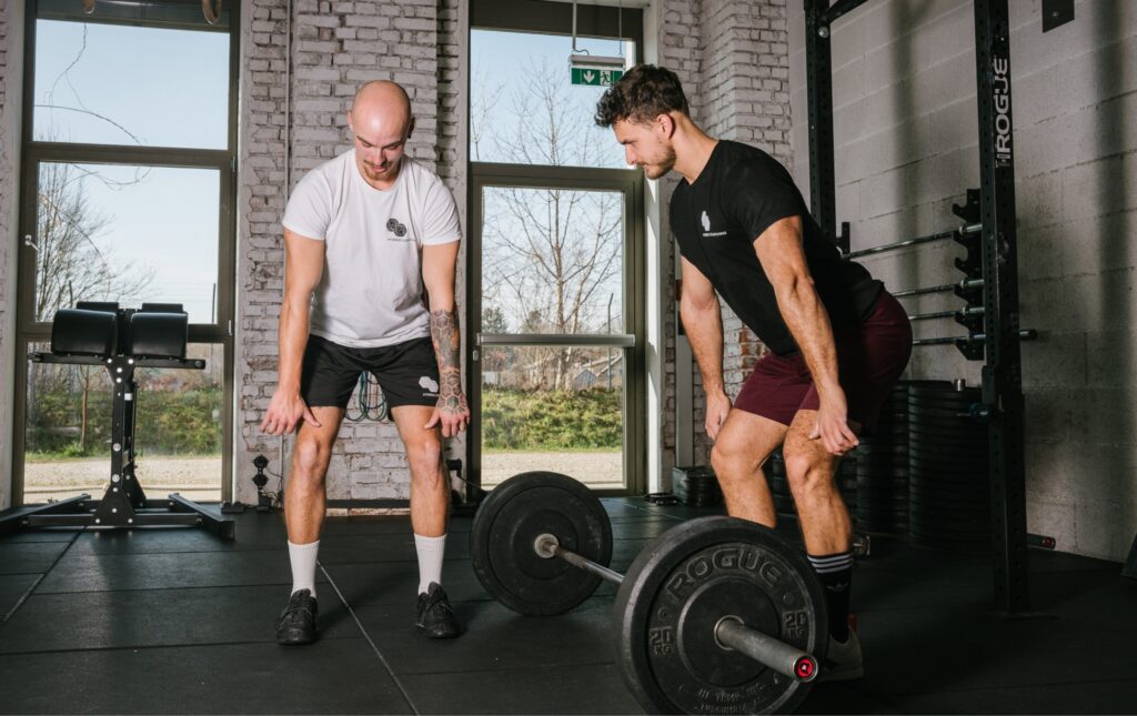 Personal Trainer zeigt Kunden Übung im Fitnessstudio vor, im Hintergrund zwei Fenster